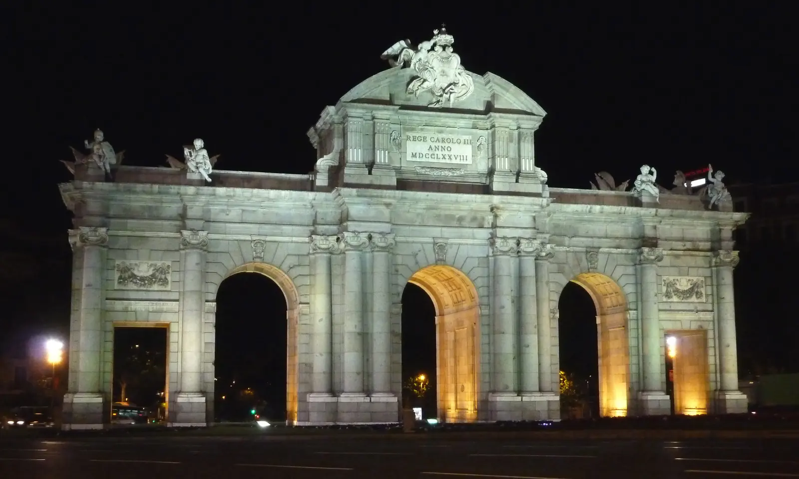 The east face of the Puerta de Alcalá, a pale stone gate with three round arches and sculpted figures above the central pediment, against a blue sky
