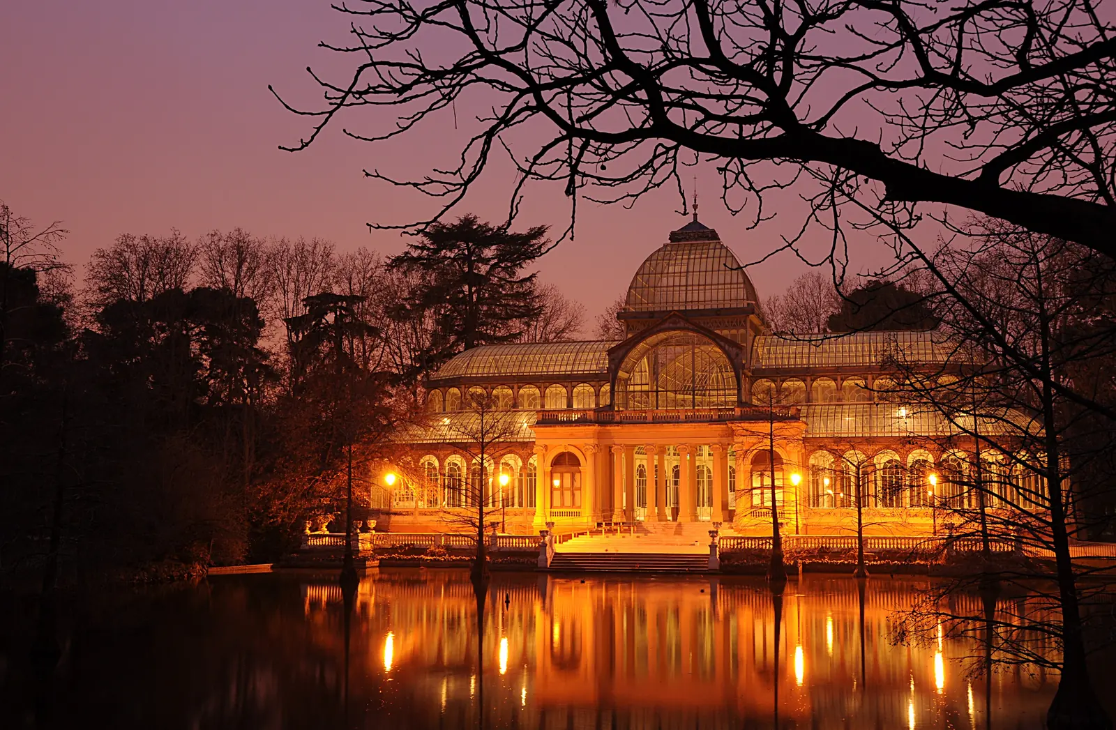 The Palacio de Cristal in Retiro Park at dusk, a glass pavilion lit orange and reflected in a still pond