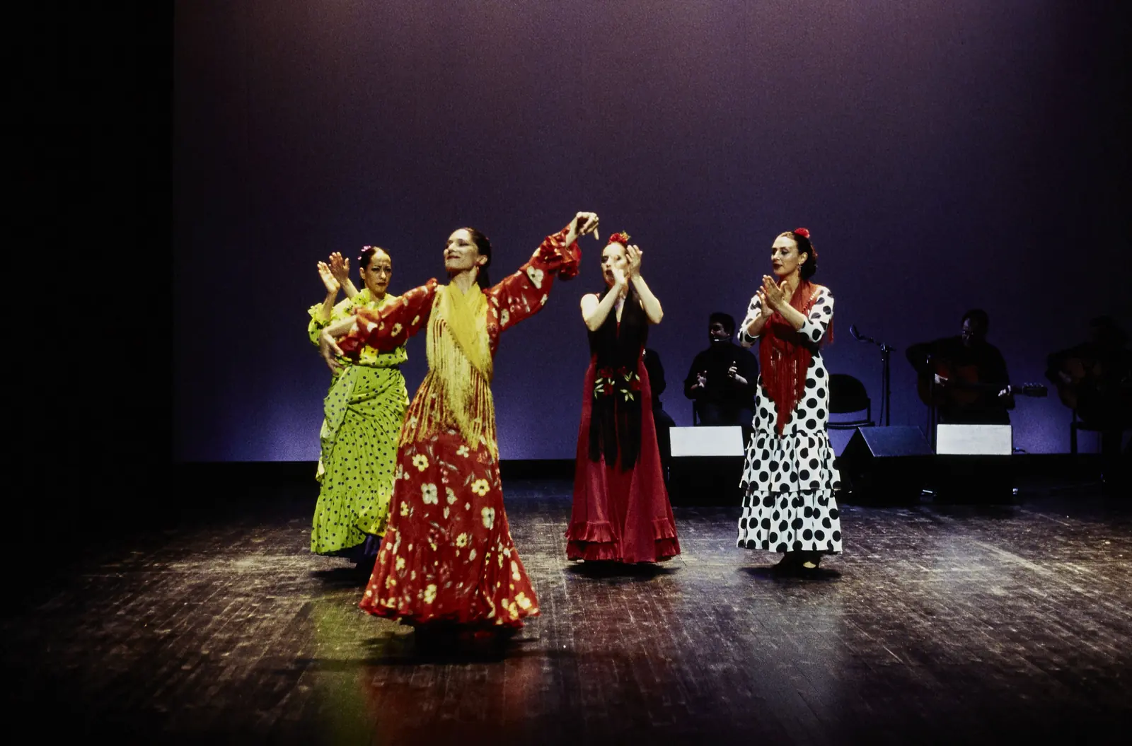 Three flamenco dancers in polka-dot and red dresses on a dark stage under violet light, with seated musicians behind them